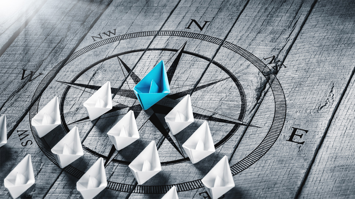 Blue Paper Boat Leading A Fleet Of Small White Boats With Compass Icon On Wooden Table